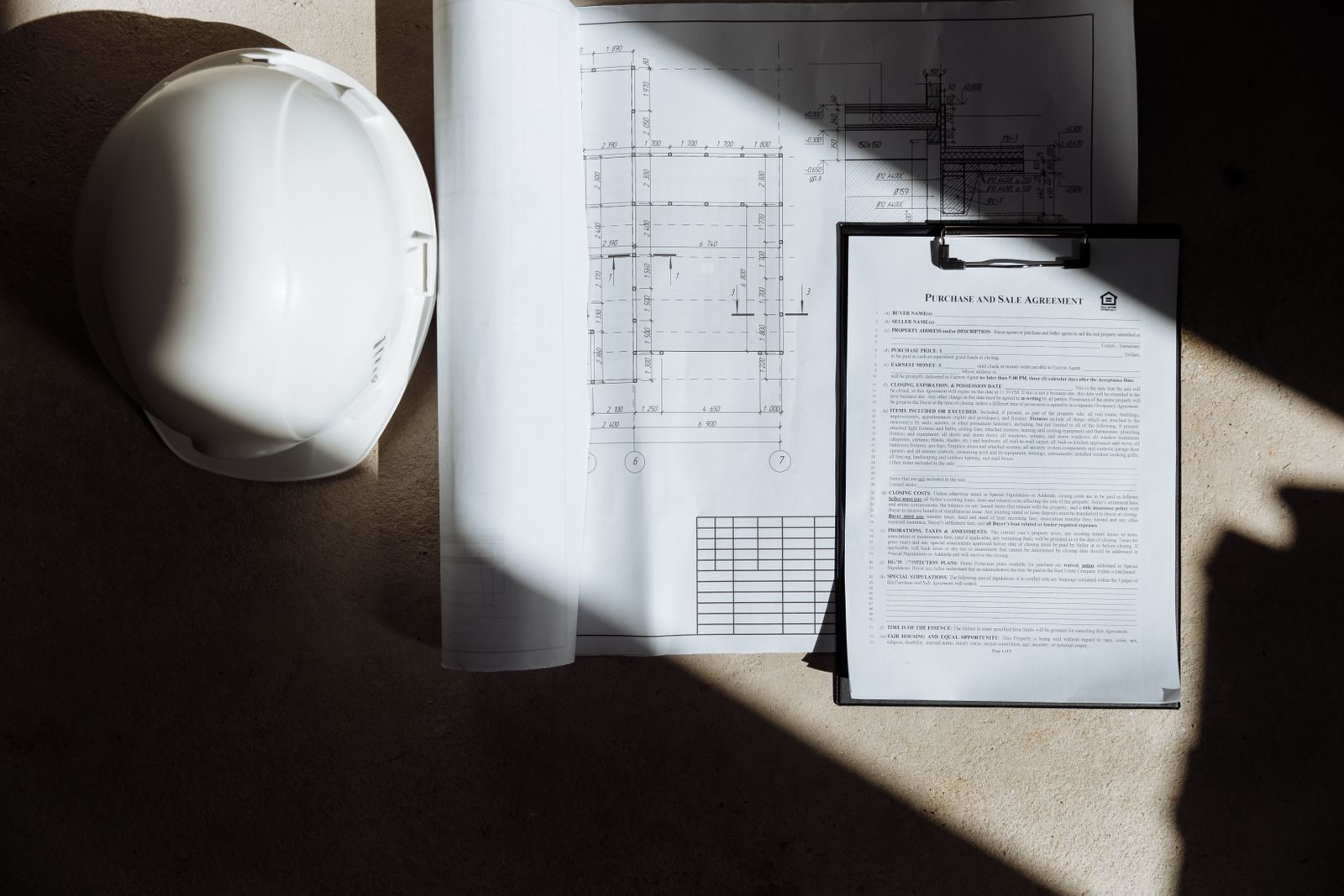 A hard hat resting beside rolled architectural blueprints and a signed agreement, in raking sunlight.