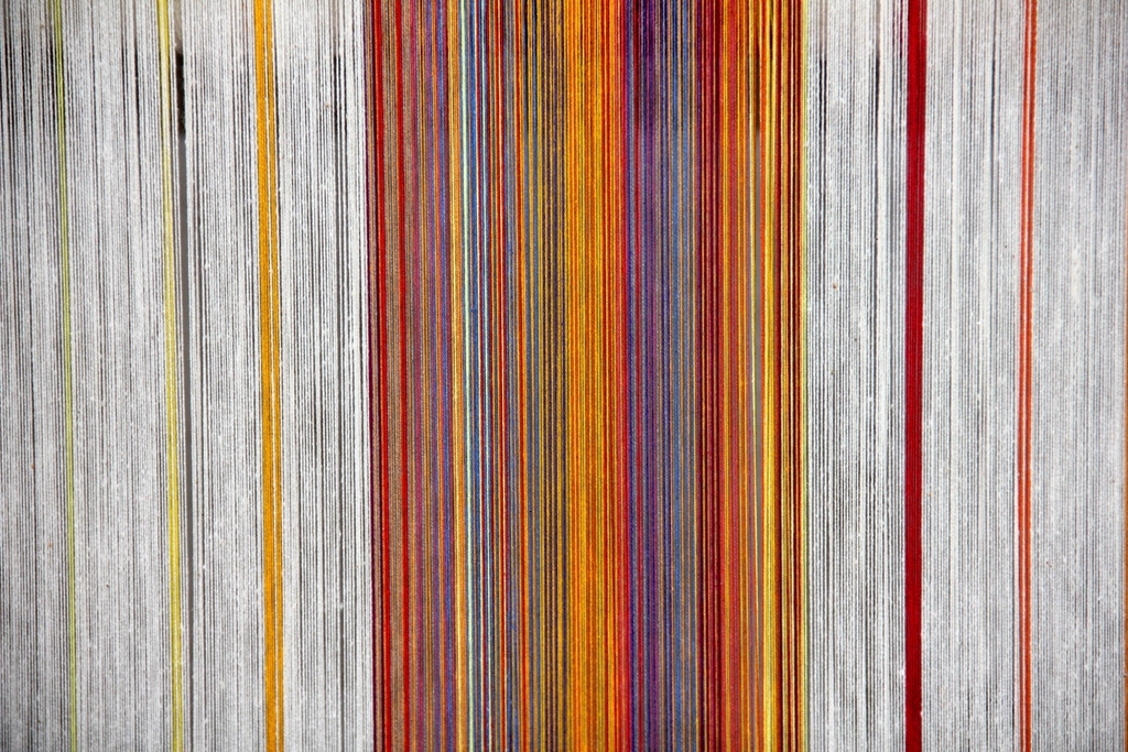 A loom warp — hundreds of parallel threads held under equal tension, coloured stripes running through undyed cotton.